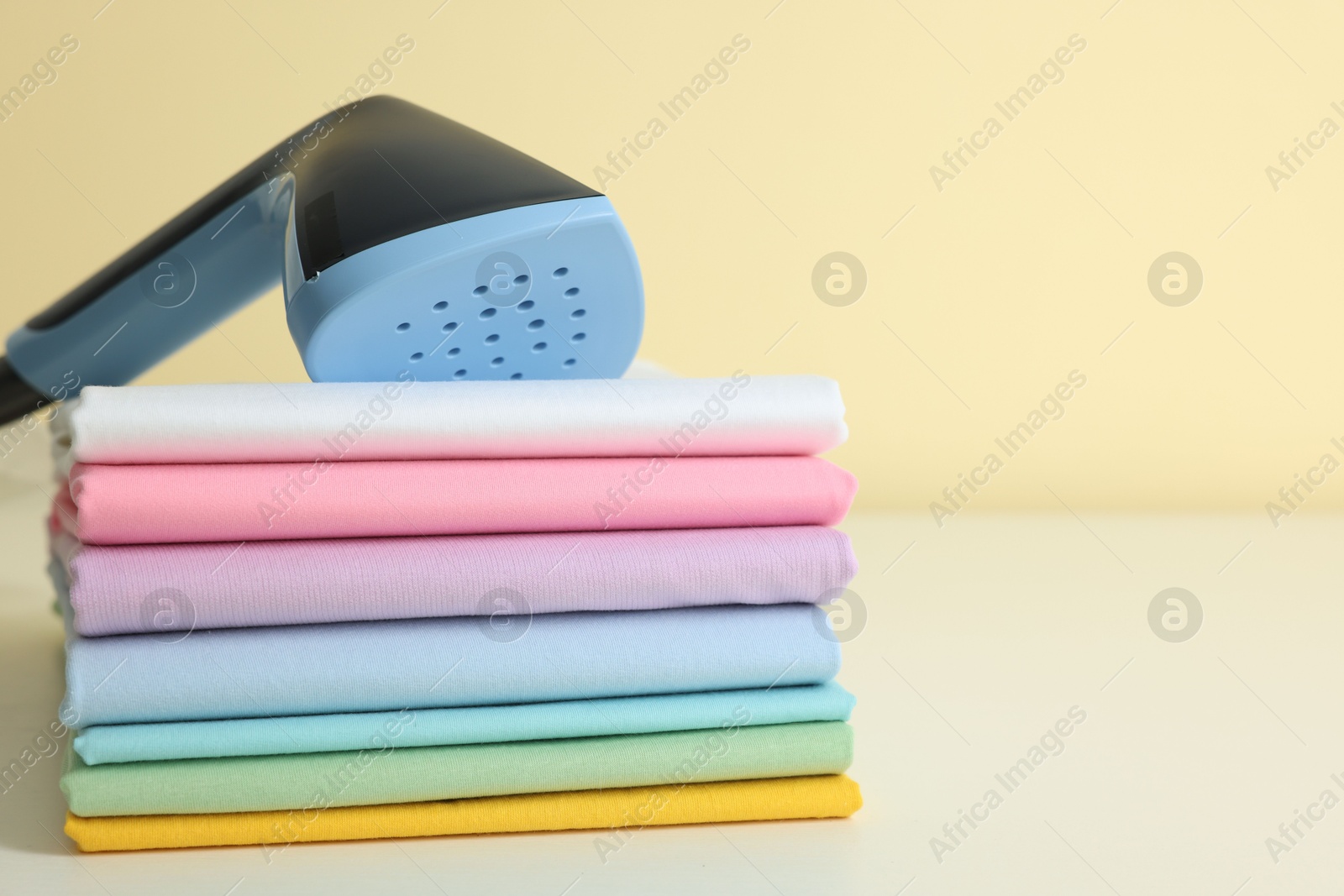 Upright steamer and stack of folded clothes on white table against beige background, closeup. Space for text Photo of Upright steamer and stack of folded clothes on white table against beige background, closeup. Space for text