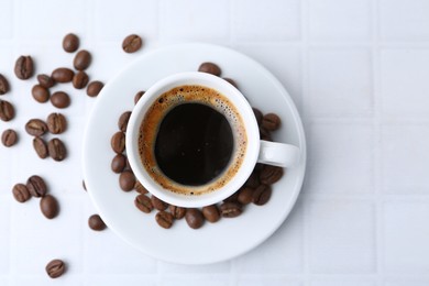 Photo of Aromatic coffee in cup and beans on white tiled table, flat lay. Space for text