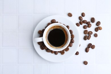 Photo of Aromatic coffee in cup and beans on white tiled table, flat lay