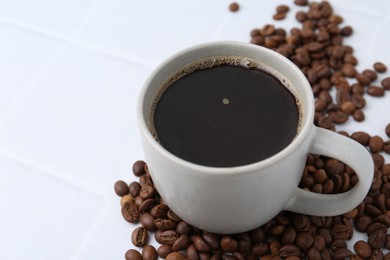 Photo of Aromatic coffee in cup and beans on white tiled table, closeup. Space for text
