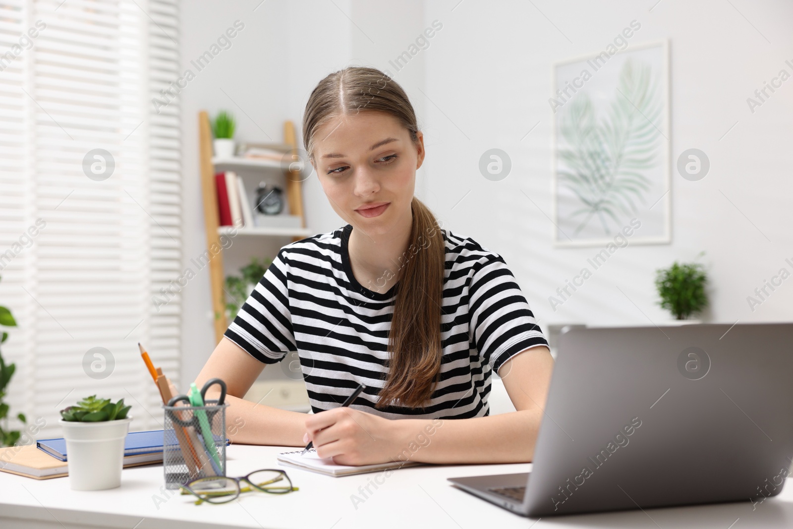 Teenage girl doing homework with laptop at white desk indoors Photo of Teenage girl doing homework with laptop at white desk indoors