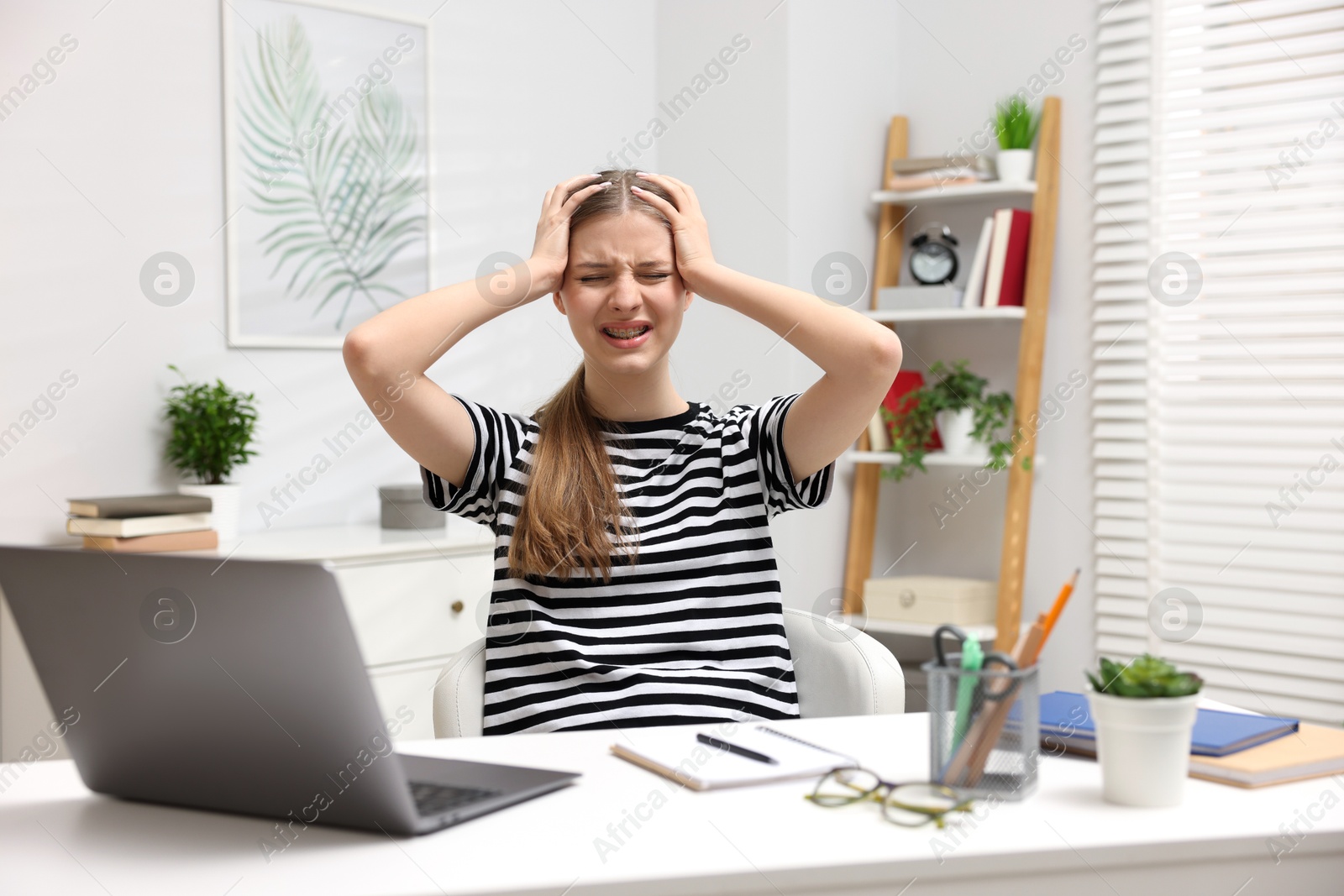 Photo of Emotional teenage girl doing homework with laptop at white desk indoors