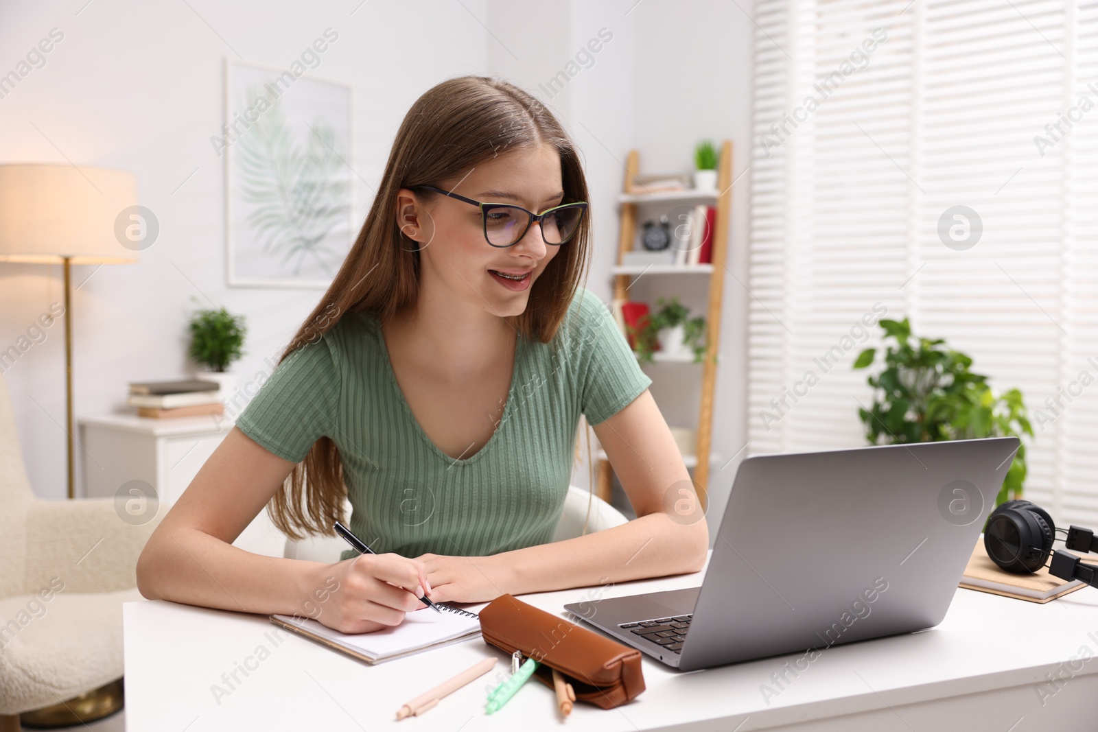Photo of Teenage girl doing homework with laptop at white desk indoors