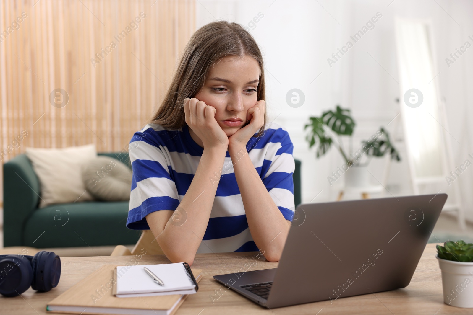 Frustrated teenage girl doing homework with laptop at wooden desk indoors Photo of Frustrated teenage girl doing homework with laptop at wooden desk indoors