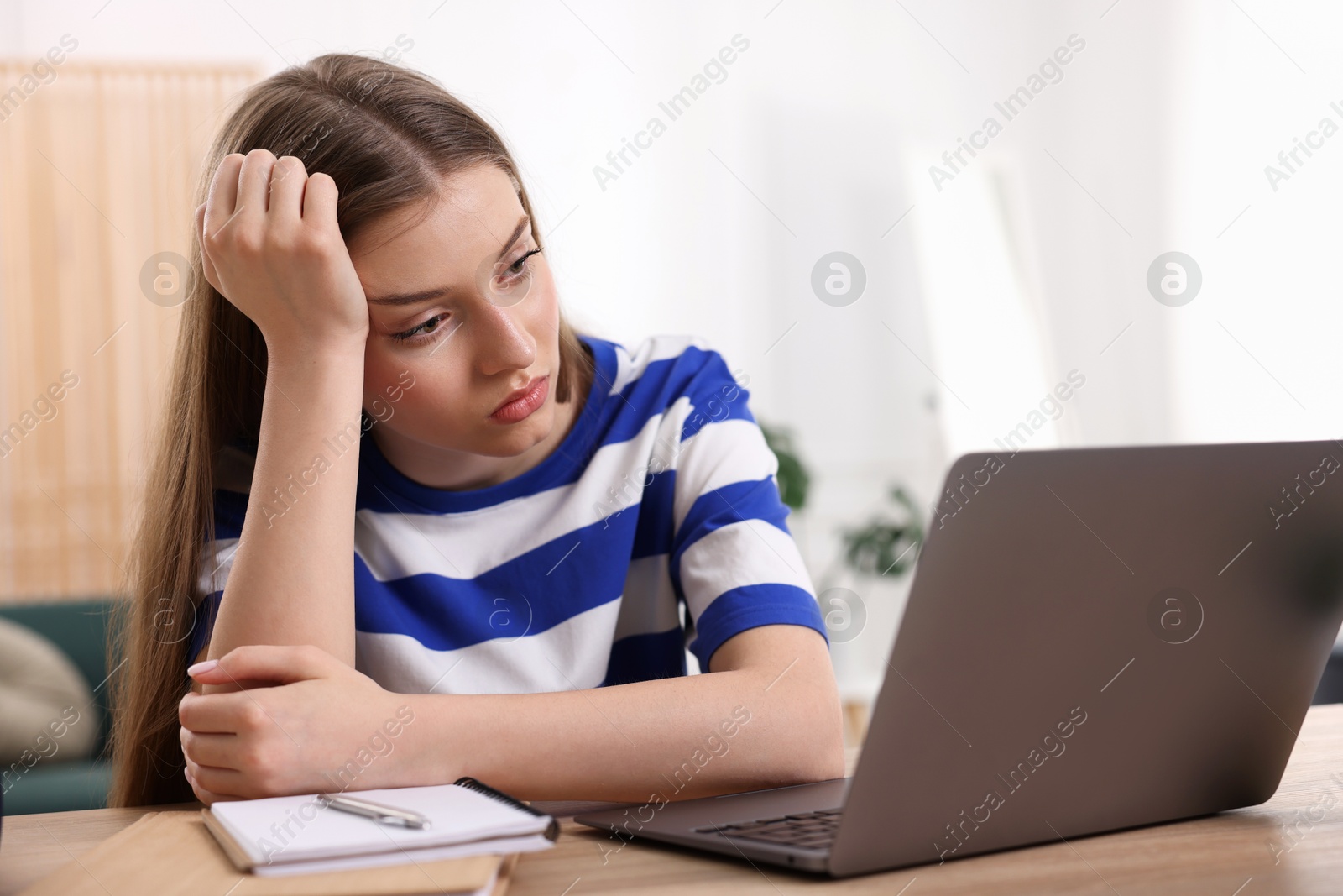 Photo of Frustrated teenage girl doing homework with laptop at wooden desk indoors