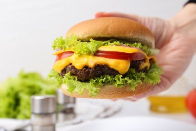 Photo of Woman holding tasty cheeseburger at white table, closeup