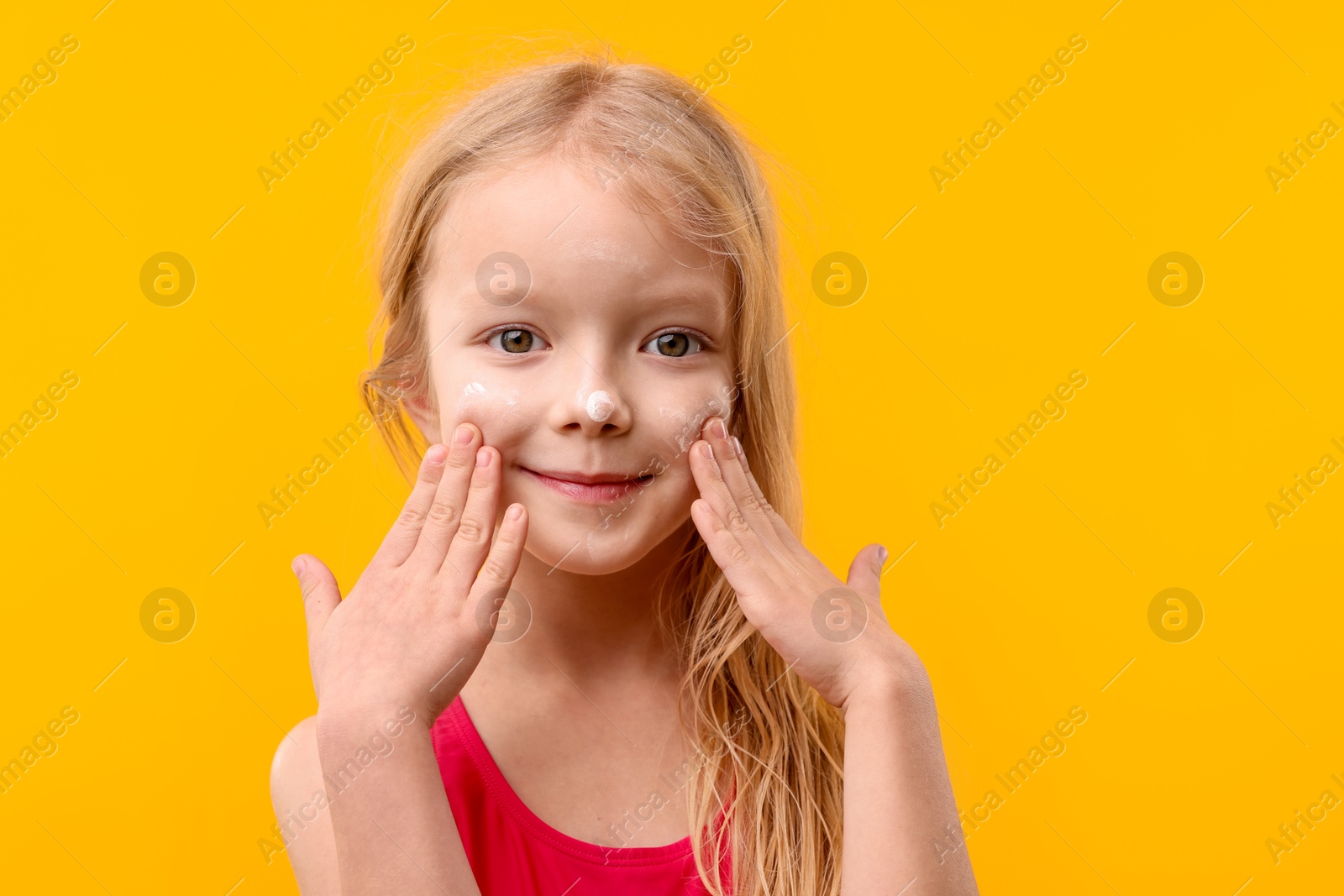 Photo of Cute little girl applying sun protection cream onto her face against yellow background, closeup