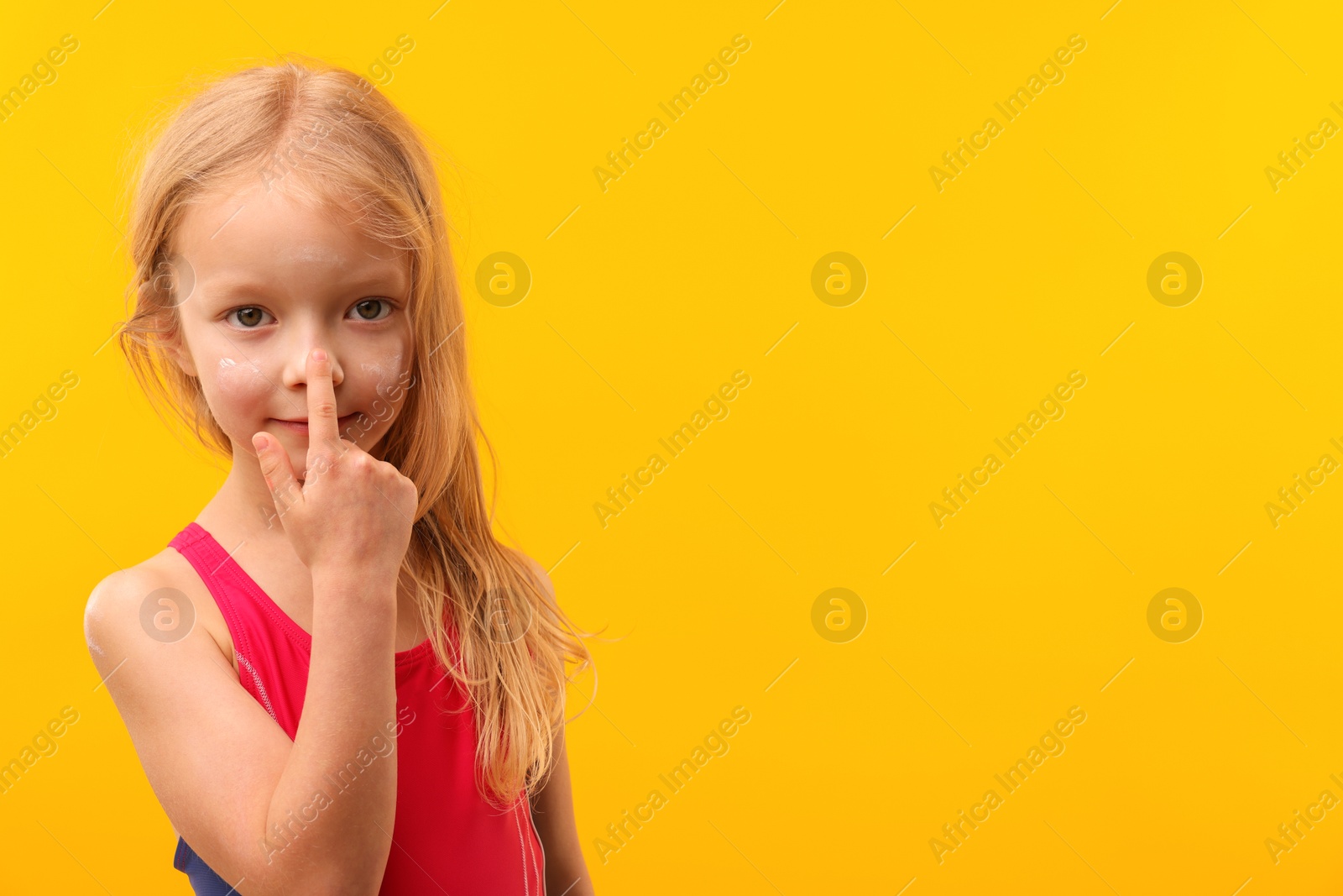 Photo of Cute little girl applying sun protection cream onto her nose against yellow background. Space for text