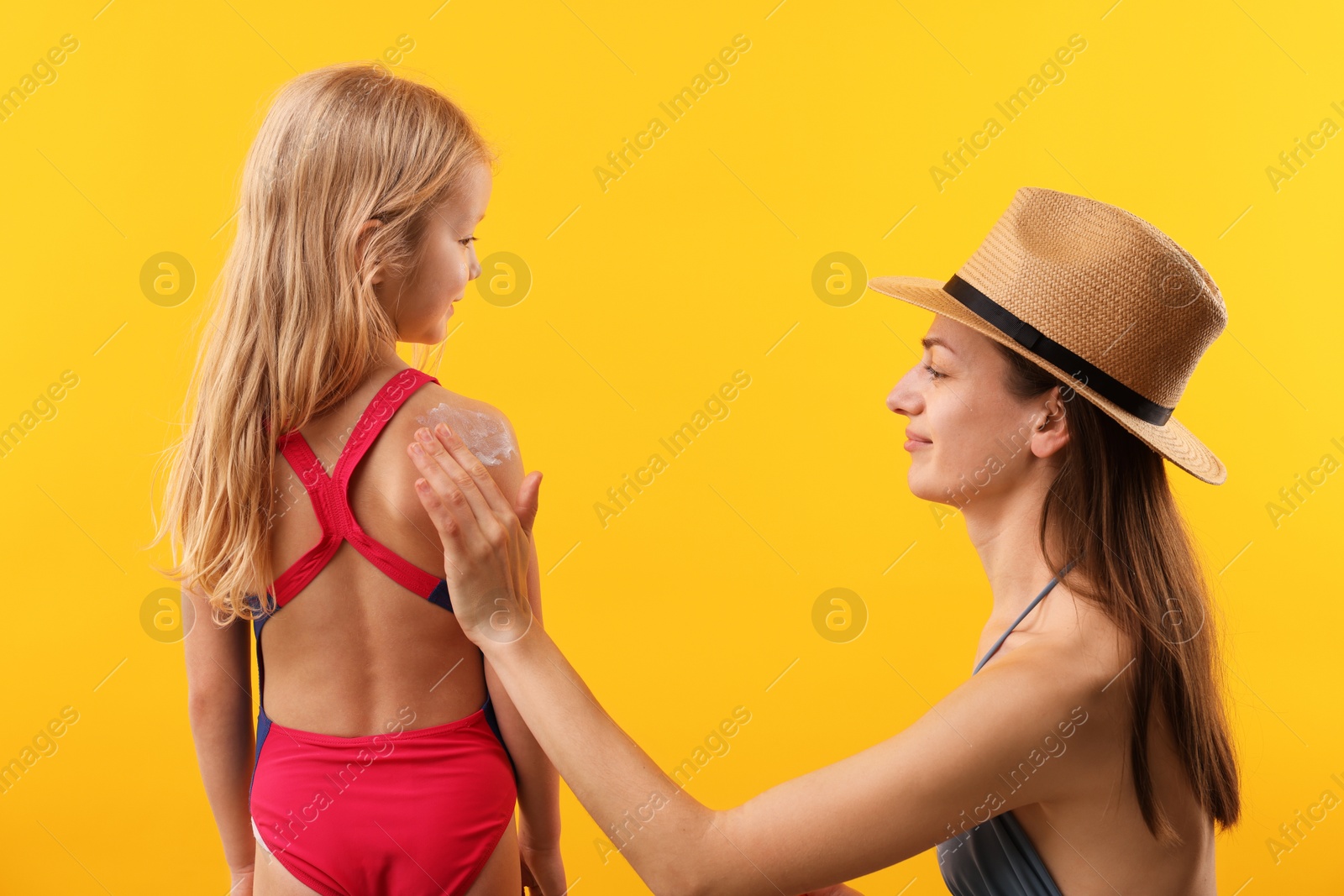 Photo of Mother applying sun protection product onto her daughter's skin on yellow background, back view