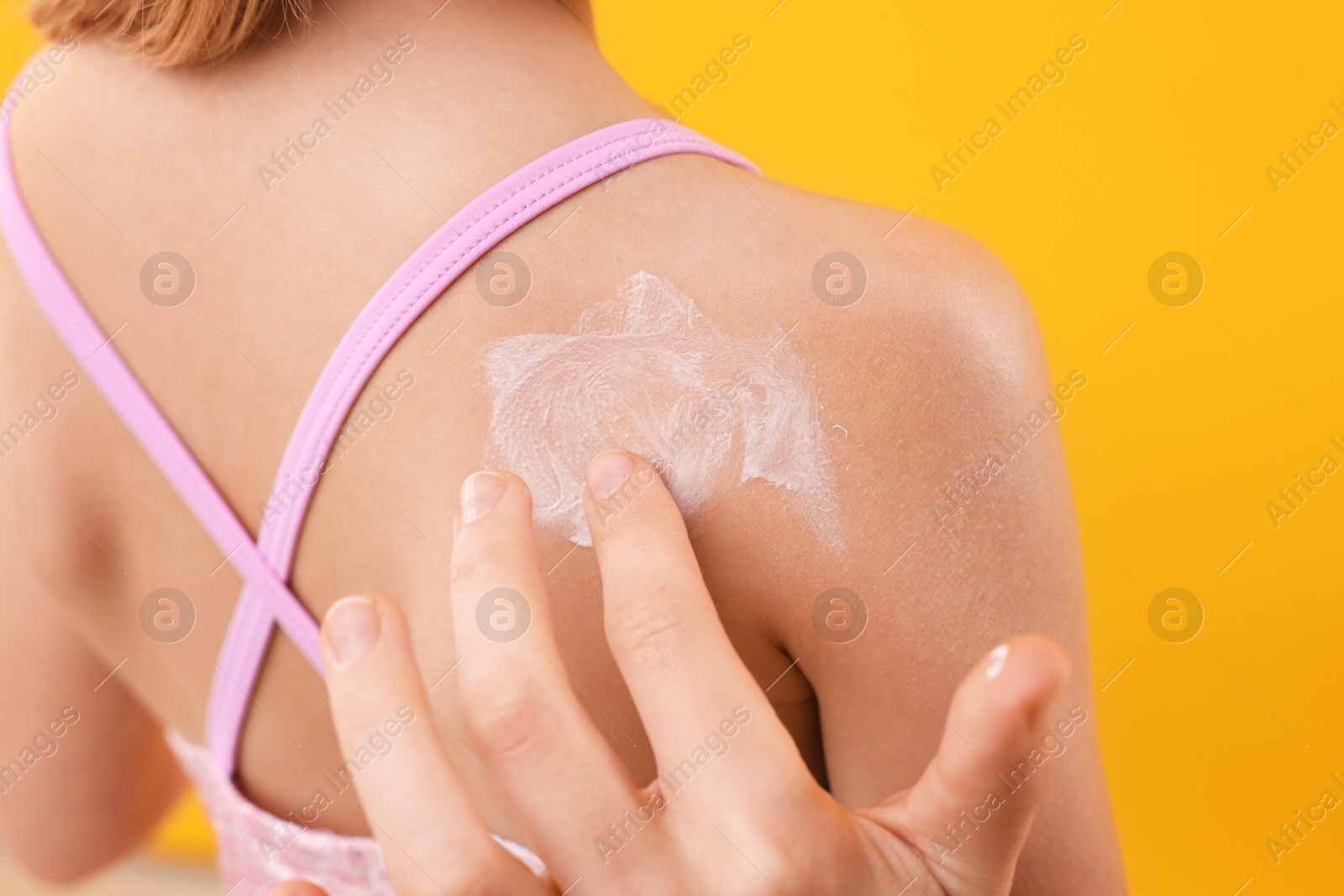 Photo of Mother applying sun protection product onto her daughter's skin on yellow background, back view