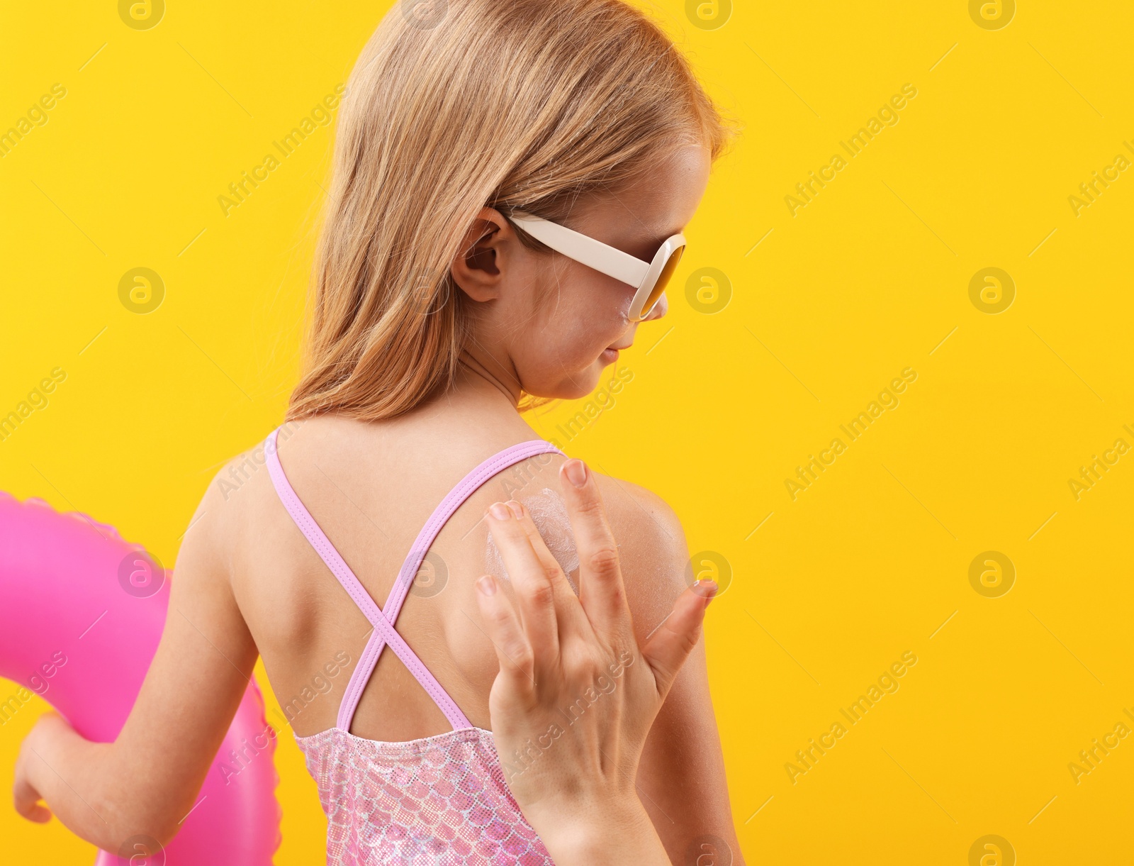 Mother applying sun protection product onto her daughter's skin on yellow background, back view Photo of Mother applying sun protection product onto her daughter's skin on yellow background, back view