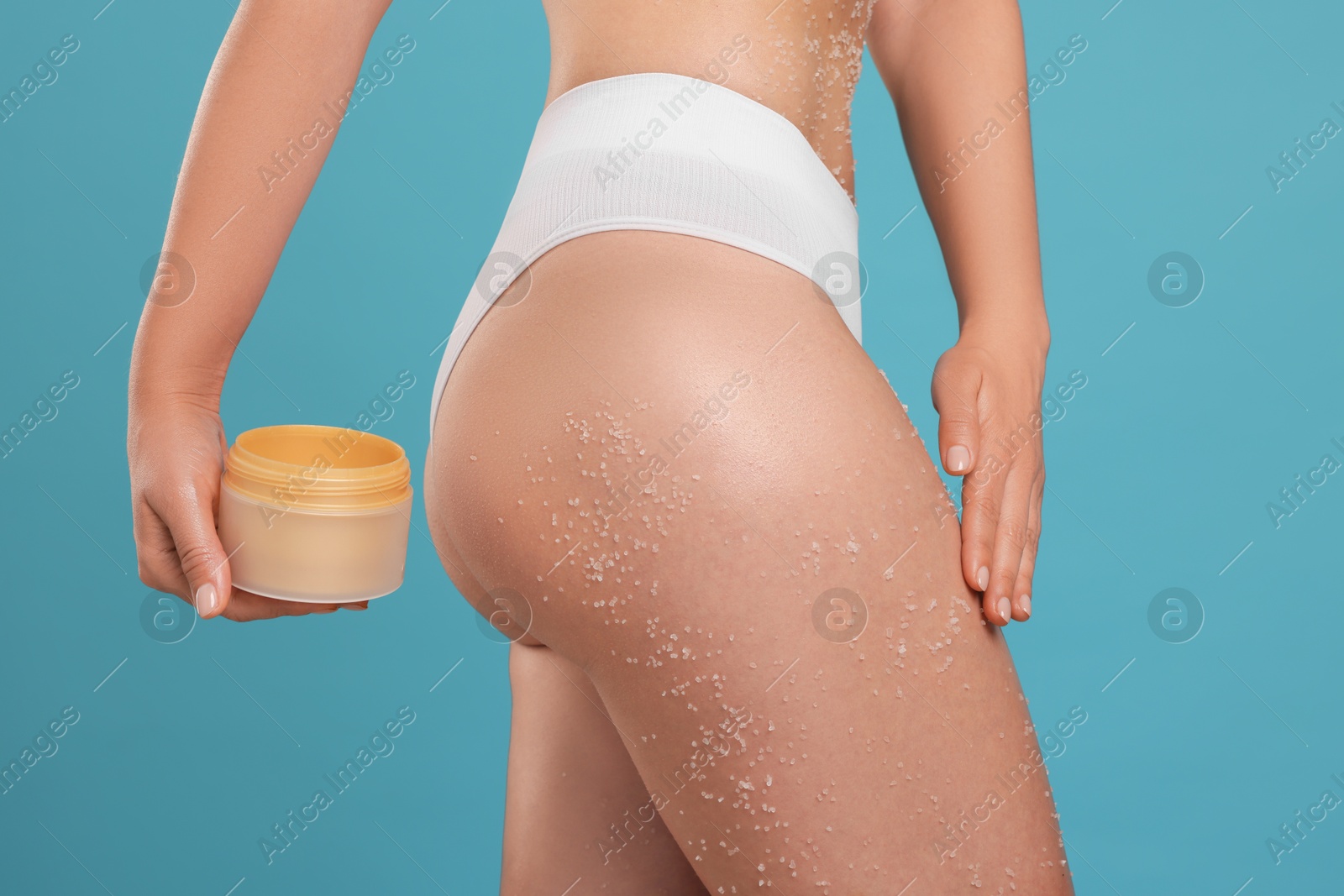 Photo of Woman with body scrub on her skin and jar against light blue background, closeup