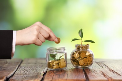 Photo of Investment concept. Woman putting coin into glass jar at wooden table against blurred green background, closeup