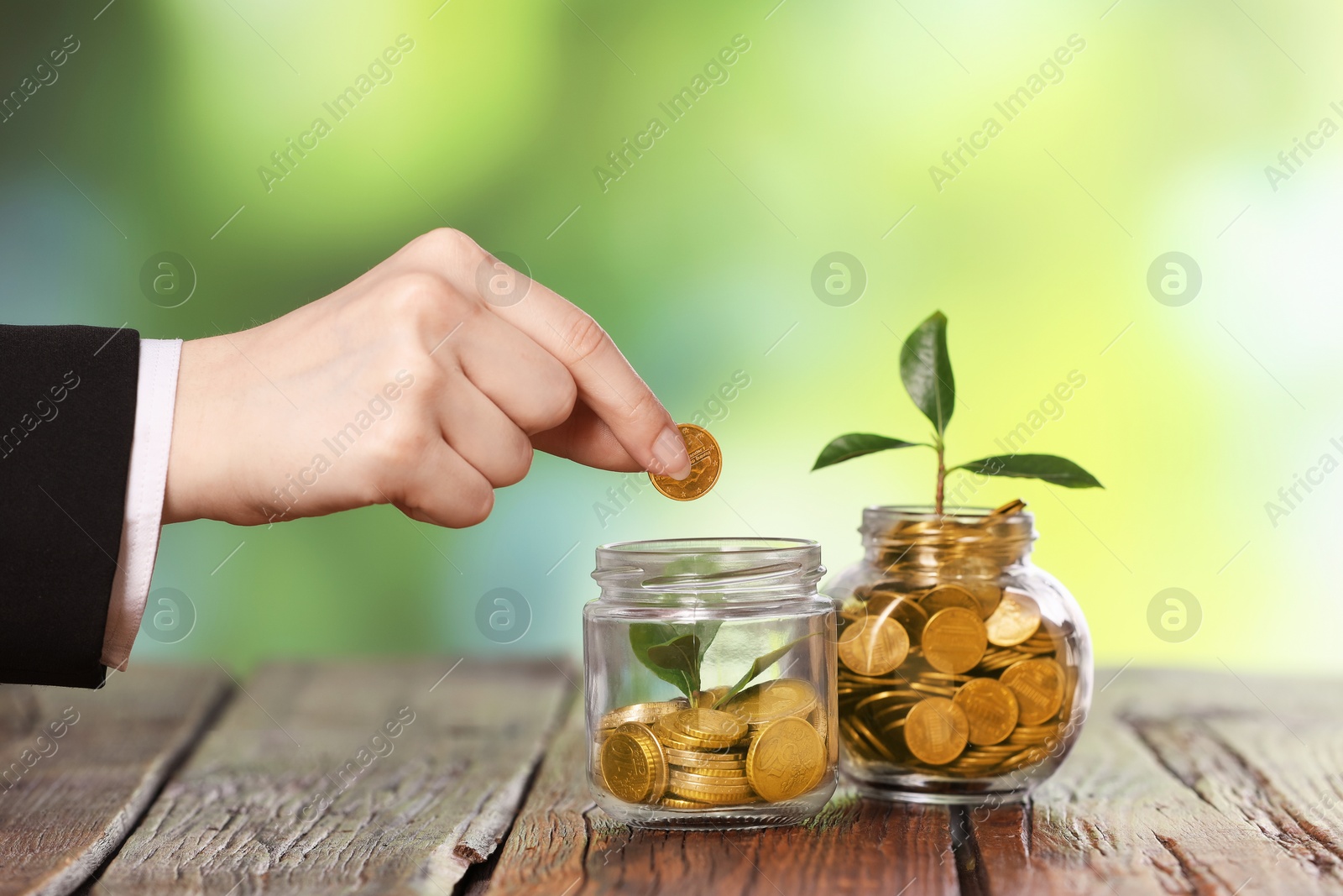 Investment concept. Woman putting coin into glass jar at wooden table against blurred green background, closeup Photo of Investment concept. Woman putting coin into glass jar at wooden table against blurred green background, closeup