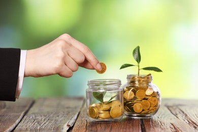 Photo of Investment concept. Woman putting coin into glass jar at wooden table against blurred green background, closeup
