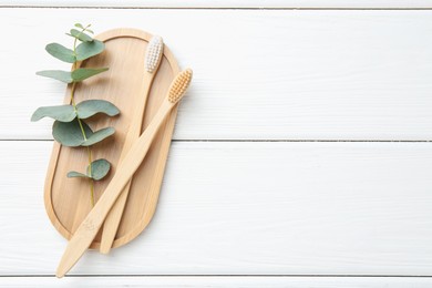 Photo of Bamboo toothbrushes and eucalyptus branch on white wooden table, top view. Space for text