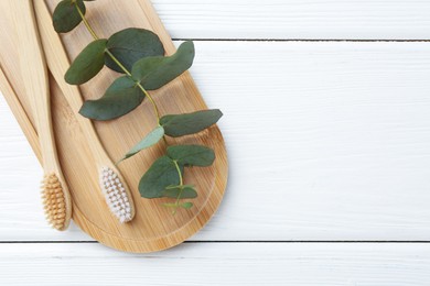 Photo of Bamboo toothbrushes and eucalyptus branch on white wooden table, top view. Space for text