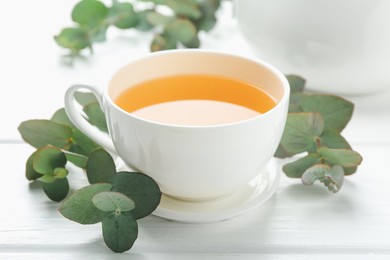 Photo of Aromatic tea in cup and eucalyptus branches on white wooden table, closeup