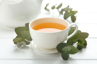 Photo of Aromatic tea in cup and eucalyptus branches on white wooden table, closeup