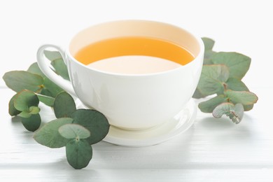 Photo of Aromatic tea in cup and eucalyptus branches on white wooden table, closeup