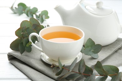 Photo of Aromatic tea in cup and eucalyptus branches on white wooden table, closeup
