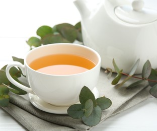 Photo of Aromatic tea in cup and eucalyptus branches on white wooden table, closeup