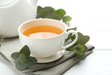 Photo of Aromatic tea in cup and eucalyptus branches on white wooden table, closeup