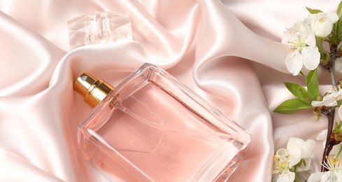 Photo of Bottle of aromatic perfume and twig with beautiful flowers on beige silk fabric, closeup