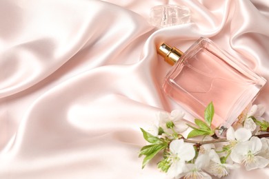 Photo of Bottle of aromatic perfume and twig with beautiful flowers on beige silk fabric, closeup. Space for text