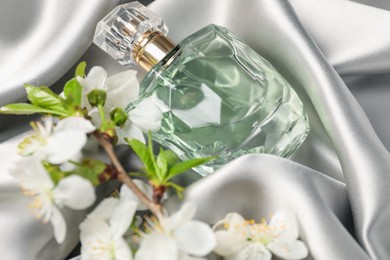 Photo of Bottle of aromatic perfume and twig with beautiful flowers on gray silk fabric, closeup