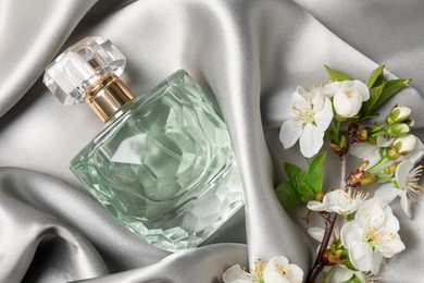Photo of Bottle of aromatic perfume and twig with beautiful flowers on gray silk fabric, flat lay