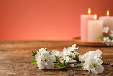 Photo of Twig with beautiful flowers and burning candles on wooden table against coral background, selective focus. Space for text