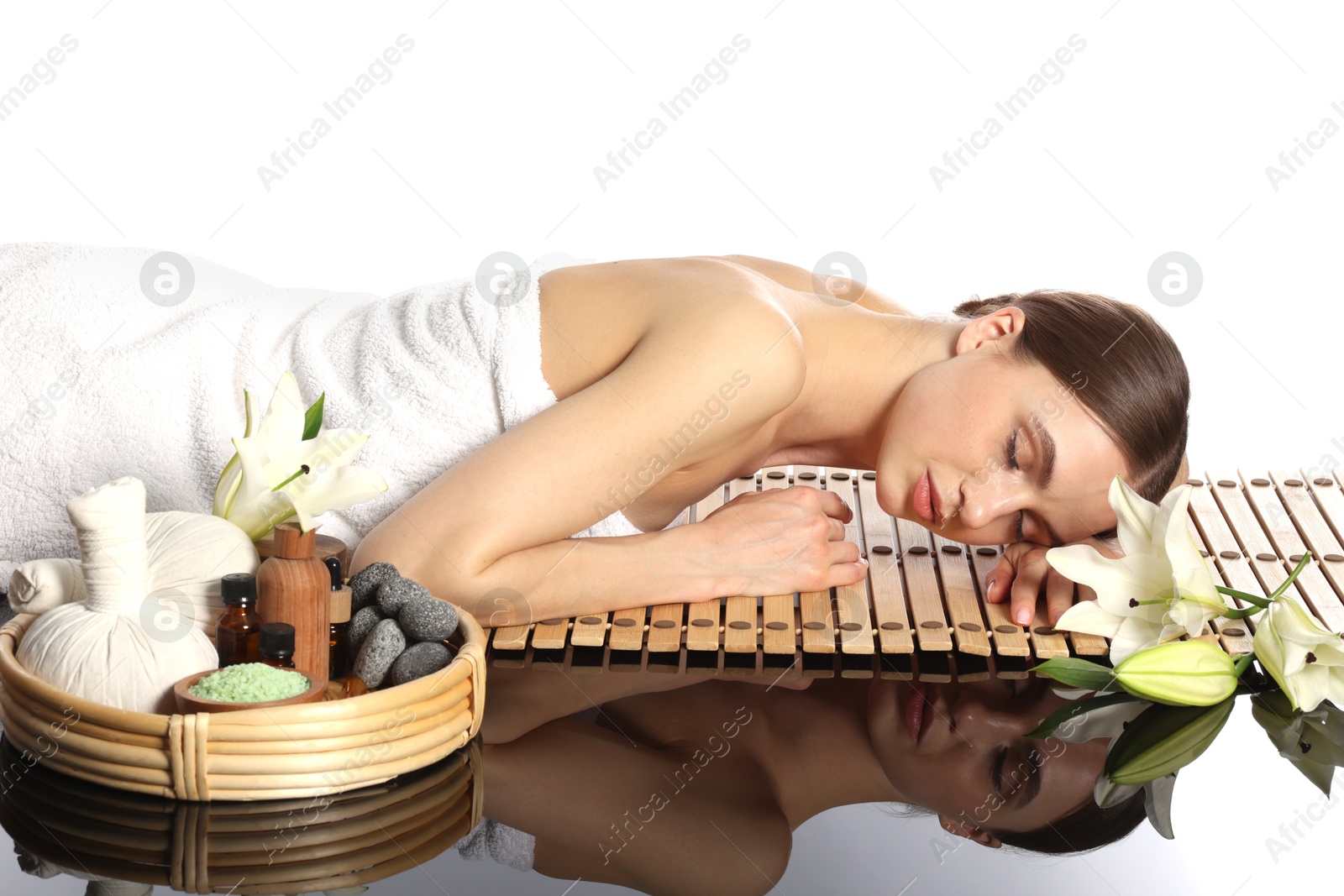 Beautiful woman lying on bamboo mat against white background. Spa products and flowers on mirror surface Photo of Beautiful woman lying on bamboo mat against white background. Spa products and flowers on mirror surface