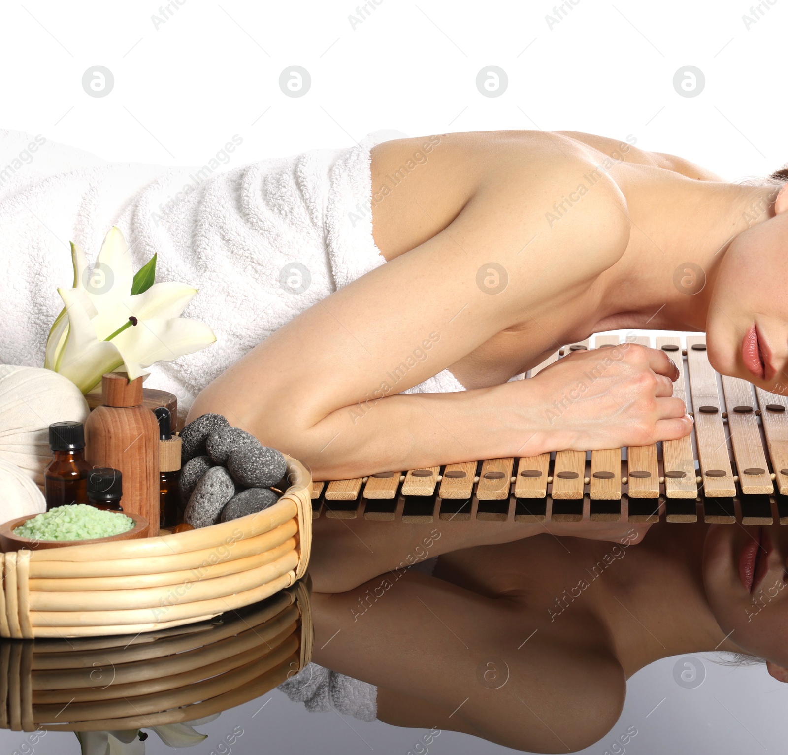 Beautiful woman lying on bamboo mat against white background. Spa products and flowers on mirror surface Photo of Beautiful woman lying on bamboo mat against white background. Spa products and flowers on mirror surface