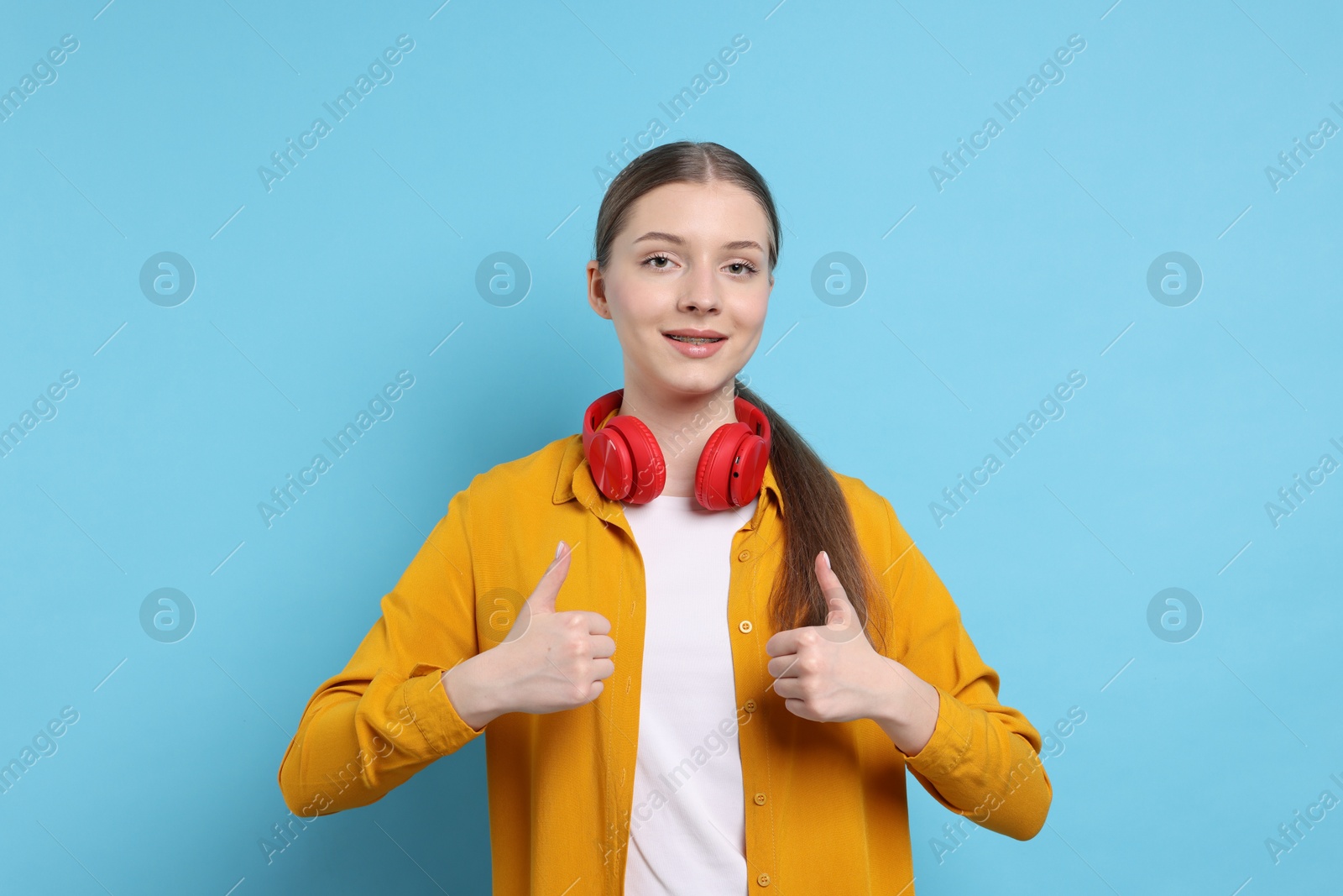 Photo of Teenage girl with headphones showing thumbs ups on light blue background