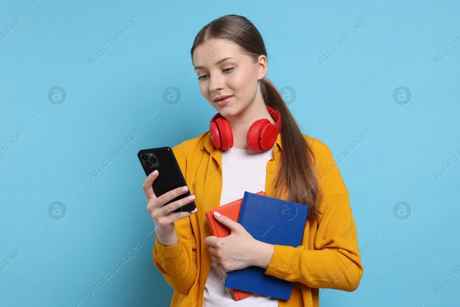 Photo of Teenage girl with headphones, smartphone and notebooks on light blue background