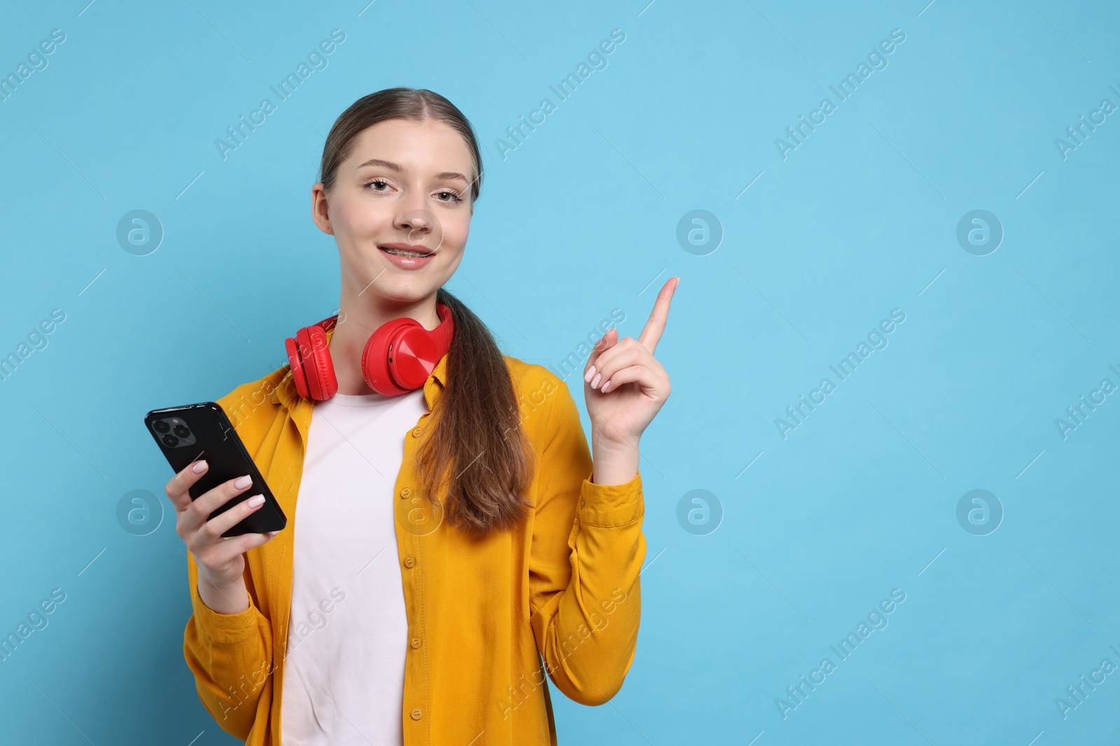 Photo of Teenage girl with headphones and smartphone pointing at something on light blue background. Space for text
