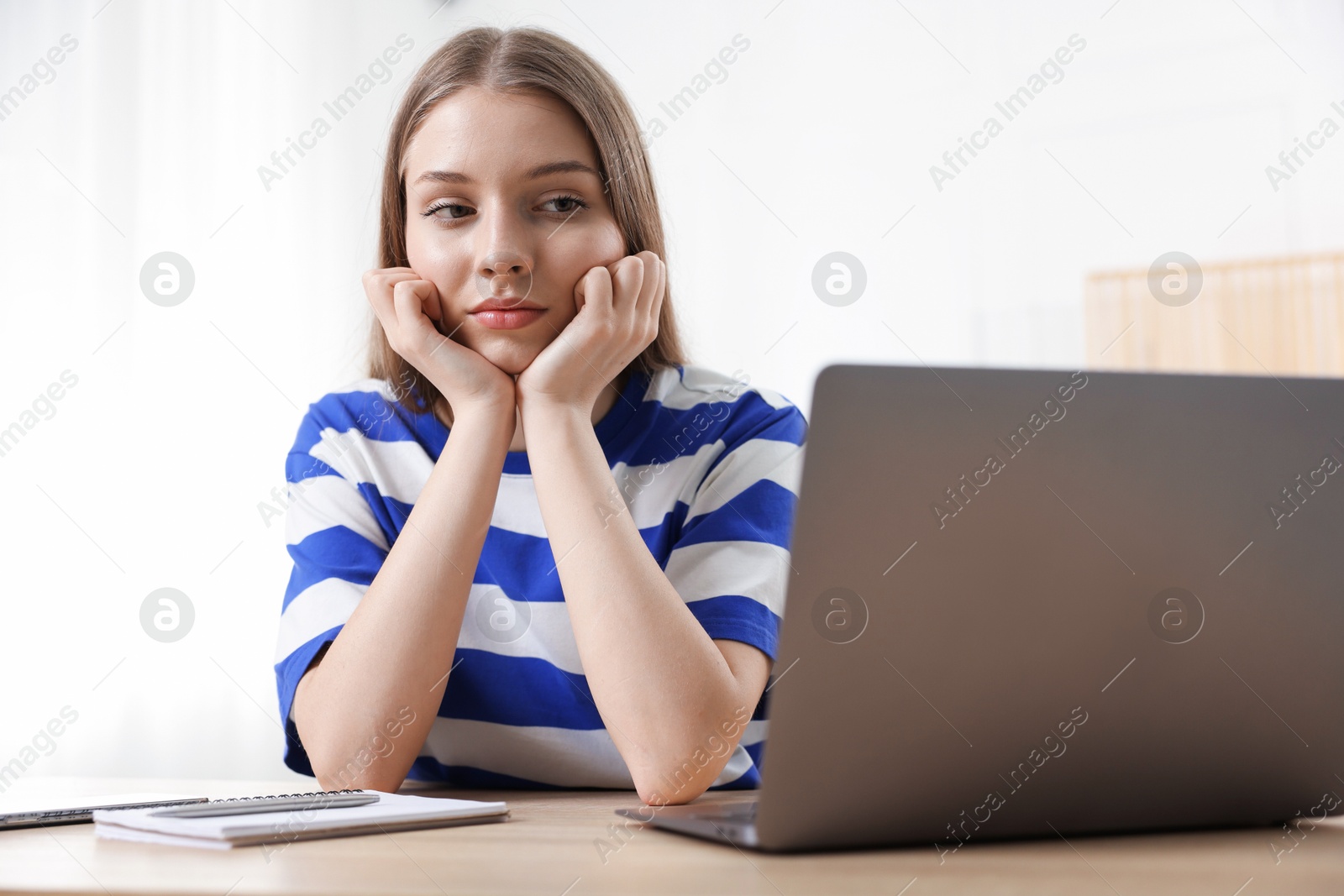 Photo of Frustrated teenage girl doing homework with laptop at wooden desk indoors