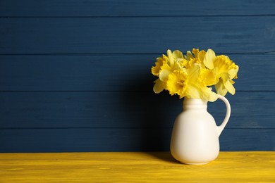 Photo of Beautiful daffodils in vase on yellow wooden table against blue background. Space for text