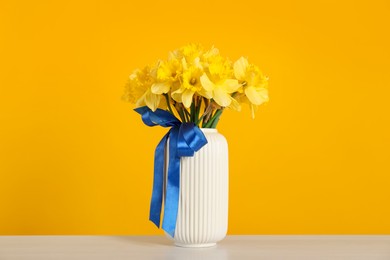 Photo of Beautiful daffodils with ribbon in vase on white table against yellow background