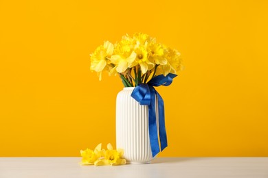 Photo of Beautiful daffodils with ribbon in vase on white table against yellow background