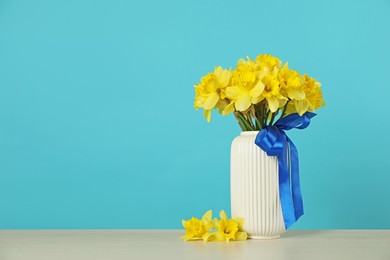 Photo of Beautiful daffodils with ribbon in vase on table against turquoise background. Space for text