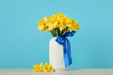 Photo of Beautiful daffodils with ribbon in vase on table against turquoise background