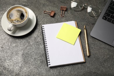 Photo of Notebook, pen, sticky note, coffee, binder clips, glasses and laptop on grey table, above view