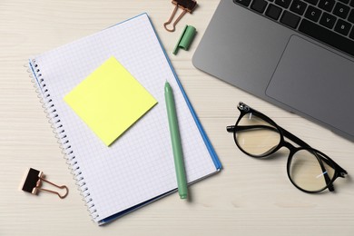 Photo of Notebook, sticky note, pen, glasses, binder clips and laptop on wooden table, flat lay