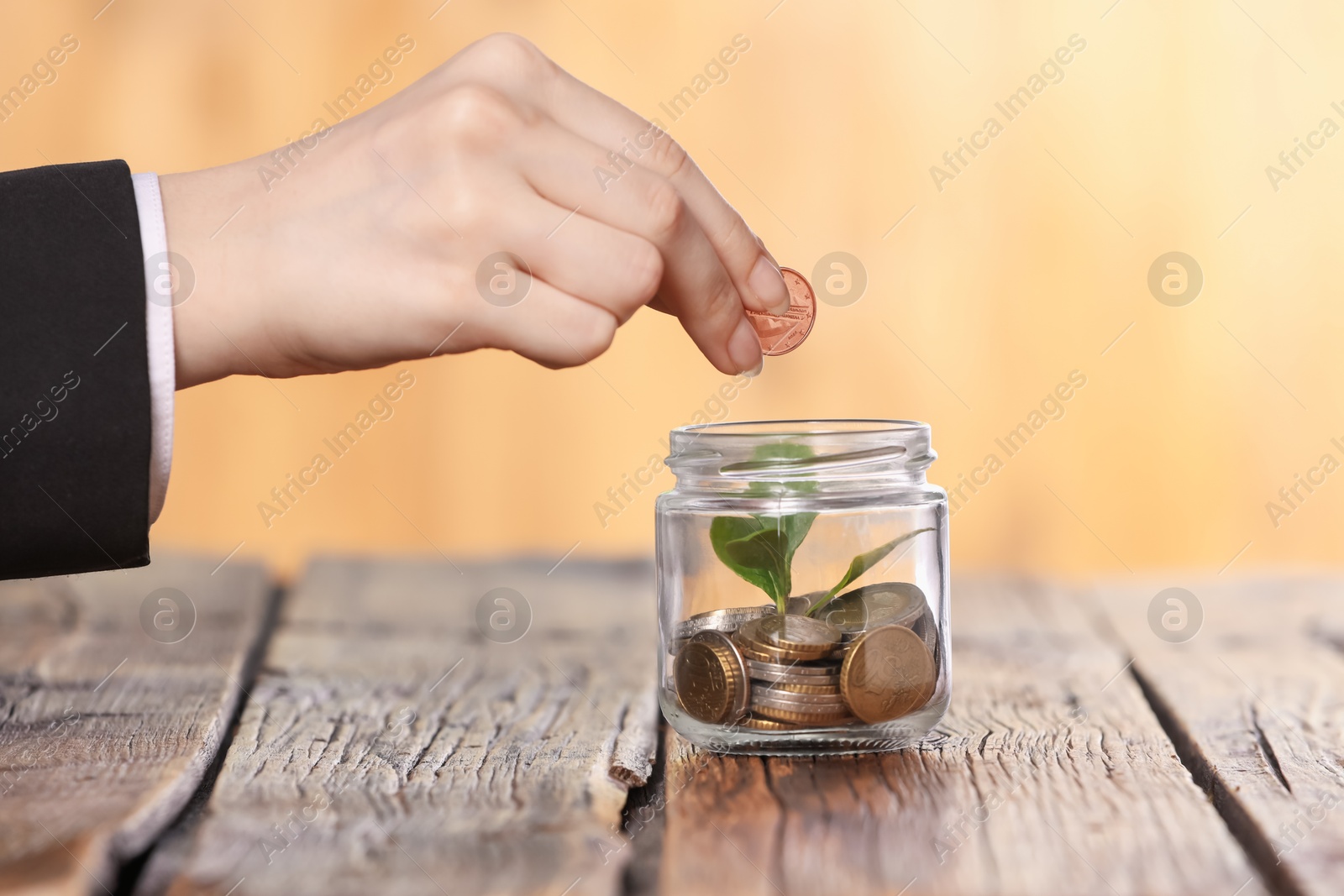 Investment concept. Woman putting coin into glass jar at wooden table against blurred orange background, closeup Photo of Investment concept. Woman putting coin into glass jar at wooden table against blurred orange background, closeup