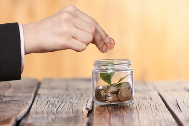 Photo of Investment concept. Woman putting coin into glass jar at wooden table against blurred orange background, closeup