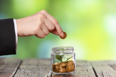 Photo of Investment concept. Woman putting coin into glass jar at wooden table against blurred green background, closeup