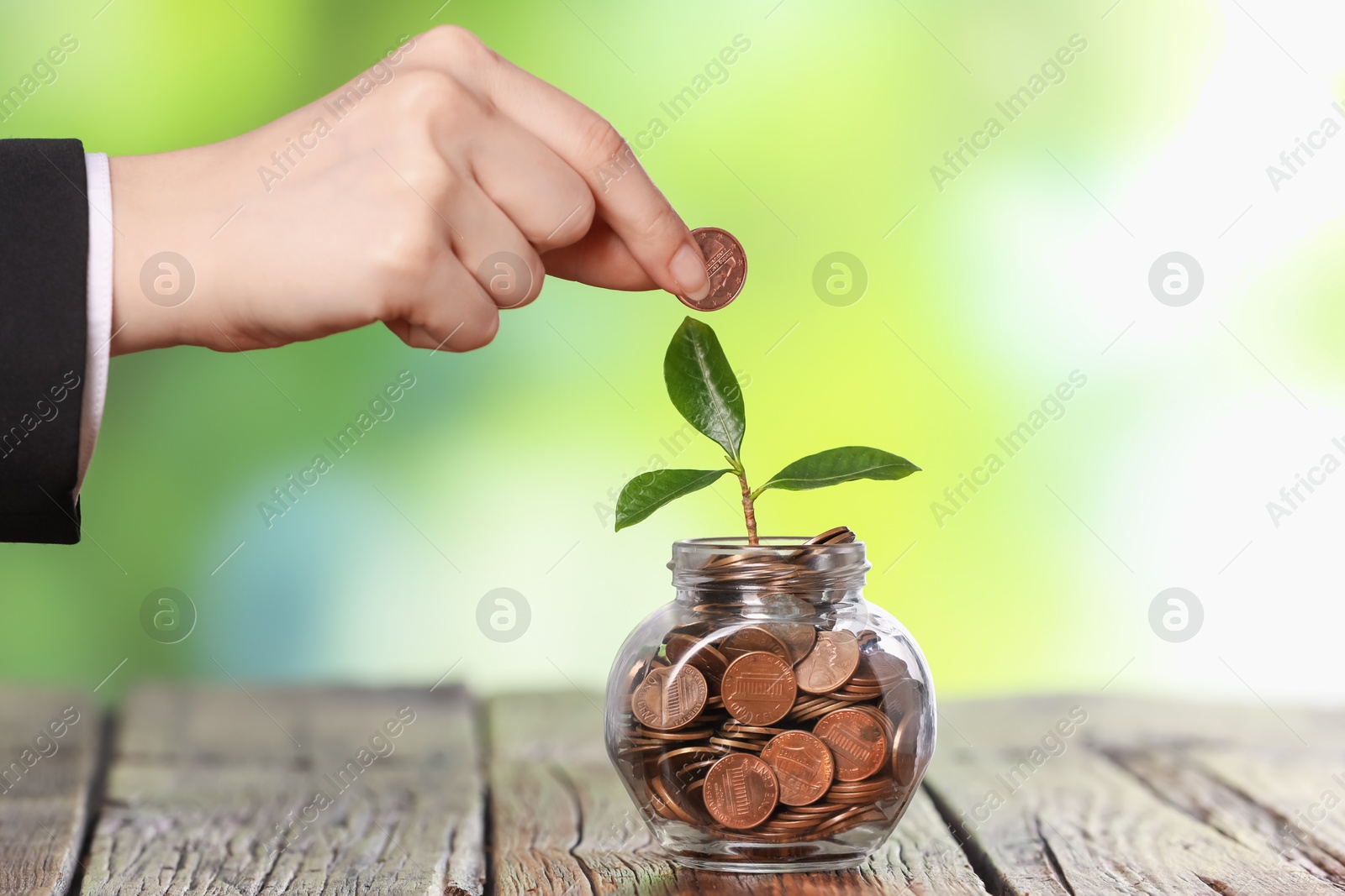 Investment concept. Woman putting coin into glass jar at wooden table against blurred green background, closeup Photo of Investment concept. Woman putting coin into glass jar at wooden table against blurred green background, closeup