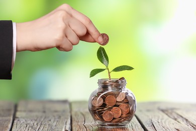 Photo of Investment concept. Woman putting coin into glass jar at wooden table against blurred green background, closeup